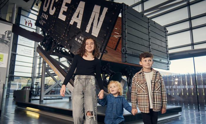 a young girl and boy hold hands with a toddler as they explore a gallery full of old engines and industrial artefacts. Behind them is a large coal tru